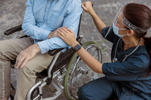 Cropped shot of nurse wearing face shield and mask holding hand, supporting mature man, recovering patient in wheelchair