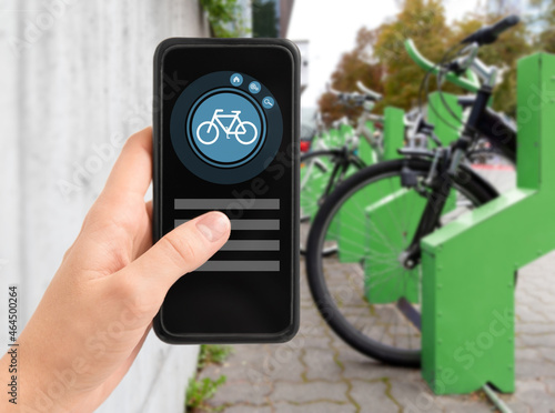 technology, transport and sustainability concept - close up of hand holding smartphone with mobile app and bicycle icon on screen over electric bike parking and charging station in city on background