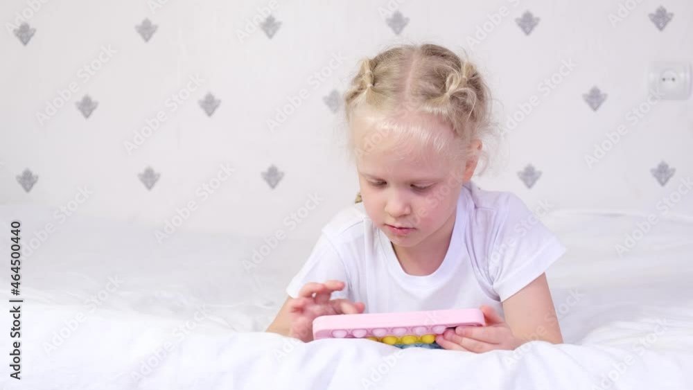 A 5-6 year old girl is lying on a bed and plays in a smartphone, pop it a smartphone case, a child and gadgets concept, the benefits and harms of a phone for a child A 5-6 year old girl is lying on a bed and plays in a smartphone, pop it a smartphone case, a child and gadgets concept, the benefits and harms of a phone for a child
