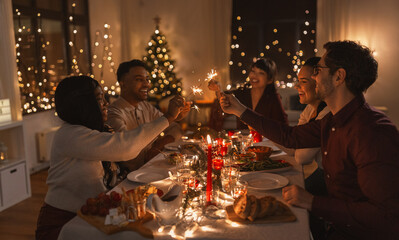 holidays and celebration concept - multiethnic group of happy friends with sparklers having christmas dinner at home