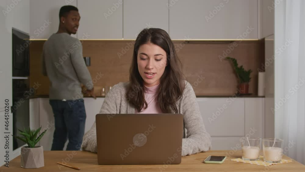Young mixed race couple discuss work on their kitchen in a bright modern apartment with a laptop and table in front Young mixed race couple discuss work on their kitchen in a bright modern apartment with a laptop and table in front