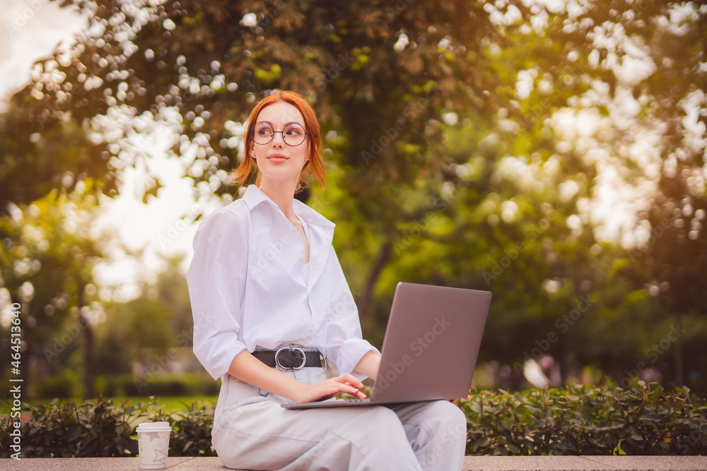 Redhead girl student in glasses and white shirt sits in the park while uses laptop with coffee to go Redhead girl student in glasses and white shirt sits in the park while uses laptop with coffee to go
