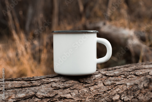 White blank metal mug mock up standing on tree outdoors, autumn forest behind. Enamelled travel cup with empty space for logo or branding