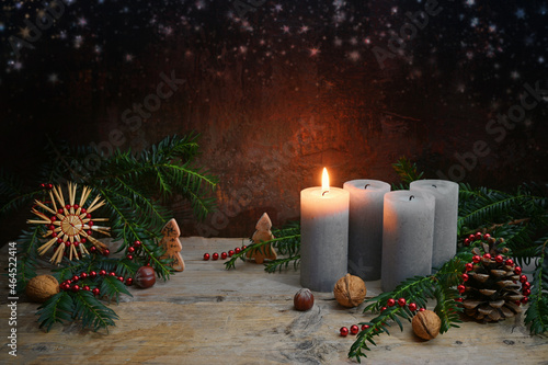 First Advent, one of four candles is lighted, Christmas decoration like nuts, straw star, cones and fir branches on rustic wood against a dark brown background, copy space