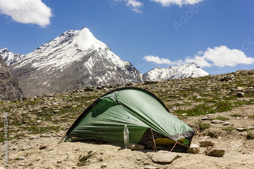 A green tent pitched on the rocky slope of a hill side with a view of snow capped Himalayan mountain peaks on a wilderness trek in Zanskar.