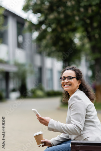 african american businesswoman with takeaway drink and mobile phone smiling at camera while sitting outdoors