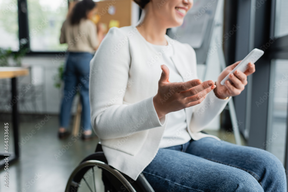cropped view of cheerful businesswoman in wheelchair gesturing while using mobile phone near blurred african american colleague