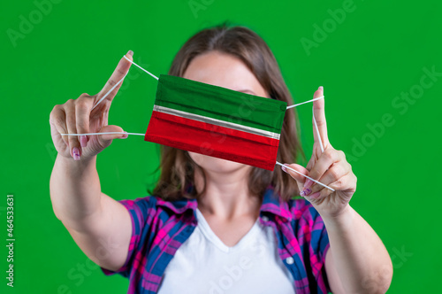 Woman in a plaid shirt holds a medical mask with of the Tatarstan flag on a green background