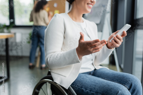 cropped view of cheerful businesswoman in wheelchair gesturing while using mobile phone near blurred african american colleague