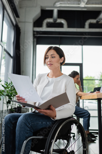 businesswoman in wheelchair working with documents near blurred african american colleague
