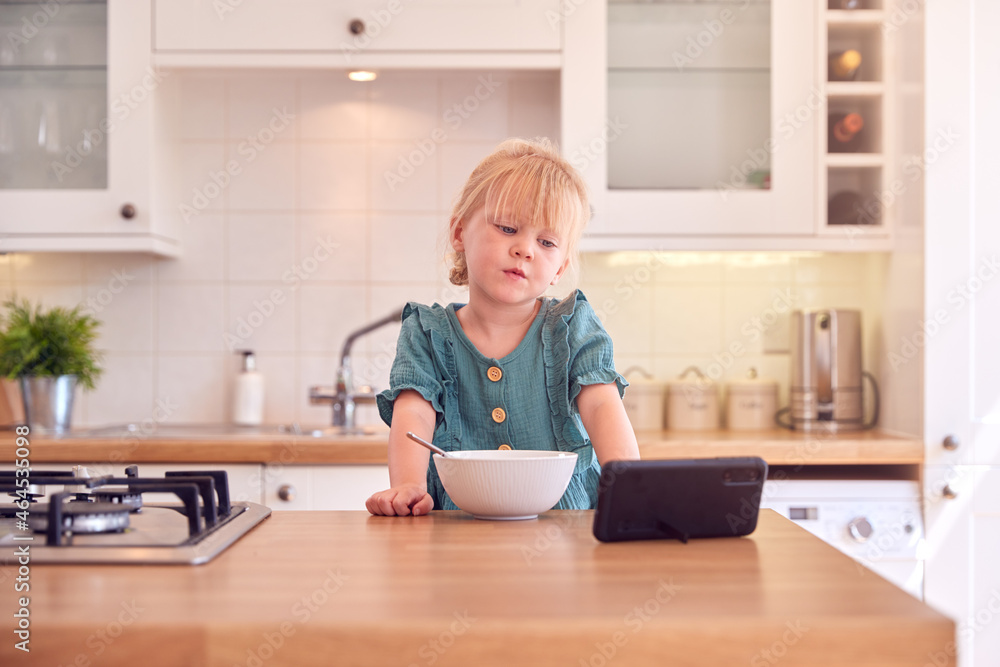 Young Girl At Home At Kitchen Counter Eating Breakfast Cereal From Bowl Whilst Watching Mobile Phone Young Girl At Home At Kitchen Counter Eating Breakfast Cereal From Bowl Whilst Watching Mobile Phone