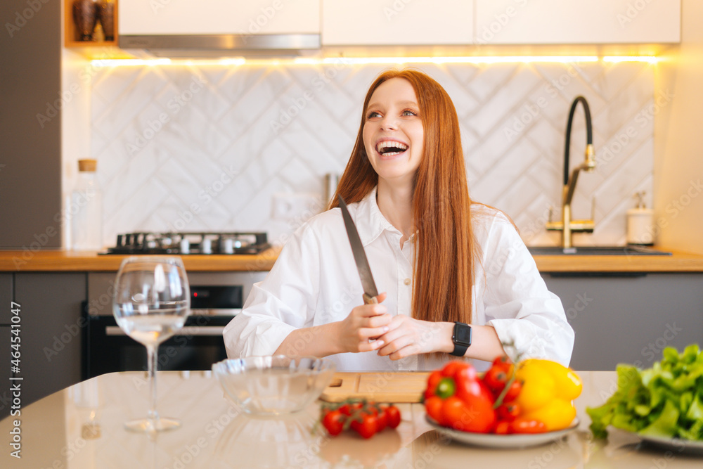 Portrait of laughing attractive redhead young woman sitting at wooden table with fresh vegetables in light kitchen room with modern interior, holding big knife, looking away.