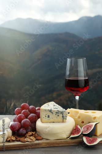 Fototapeta Naklejka Na Ścianę i Meble -  Different types of delicious cheeses, fruits and wine on wooden table against mountain landscape