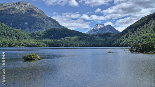 A wide view of rocks in the calm water of Lake Sylvan, thick green forest against a background of snowcapped Mount Earnslaw and cloudy blue sky, South Island, New Zealand