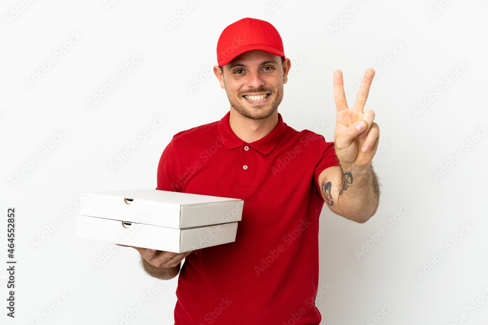 pizza delivery man with work uniform picking up pizza boxes over isolated white wall smiling and showing victory sign pizza delivery man with work uniform picking up pizza boxes over isolated white wall smiling and showing victory sign