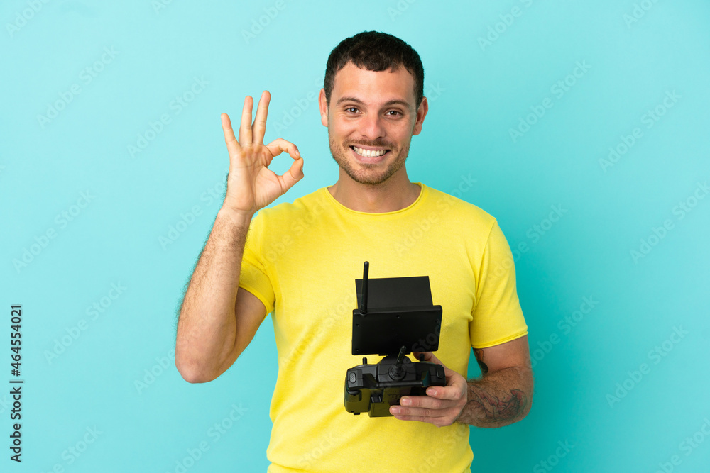 Brazilian man holding a drone remote control over isolated blue background showing ok sign with fingers Brazilian man holding a drone remote control over isolated blue background showing ok sign with fingers