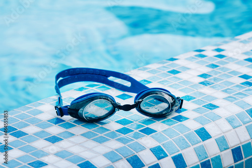 Blue professional swimming goggles with water drops on yellow lenses on a blue and white tiles of pool floor.