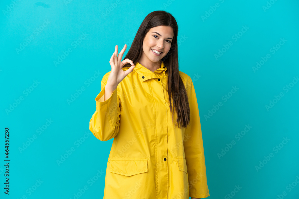 Teenager Brazilian girl wearing a rainproof coat over isolated blue background showing ok sign with fingers Teenager Brazilian girl wearing a rainproof coat over isolated blue background showing ok sign with fingers