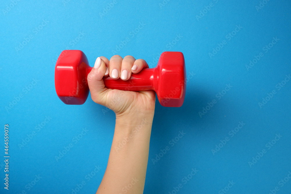 Sports concept. A female hand holds a red plastic dumbbell. Blue background. Hand of woman lifting dumbbell