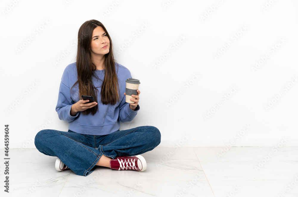 Young woman sitting on the floor holding coffee to take away and a mobile while thinking something Young woman sitting on the floor holding coffee to take away and a mobile while thinking something