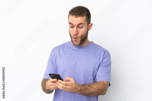 Young handsome caucasian man isolated on white background looking at the camera while using the mobile with surprised expression