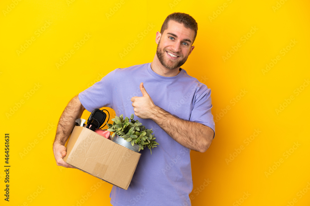Young caucasian making a move while picking up a box full of things isolated on yellow background giving a thumbs up gesture Young caucasian making a move while picking up a box full of things isolated on yellow background giving a thumbs up gesture