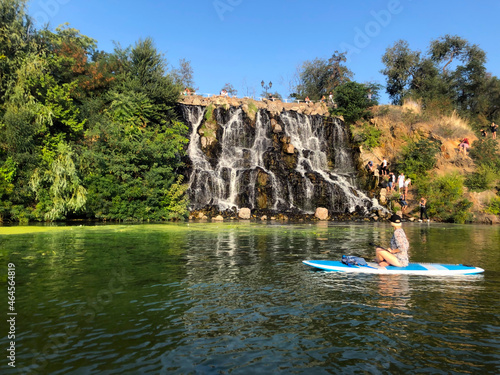 Monastyrsky Island, Dnipro, Ukraine. A group of tourists descends to artificial waterfall. A girl on sup rafting down the river. The waterfall is surrounded by trees against a blue sky without clouds photo