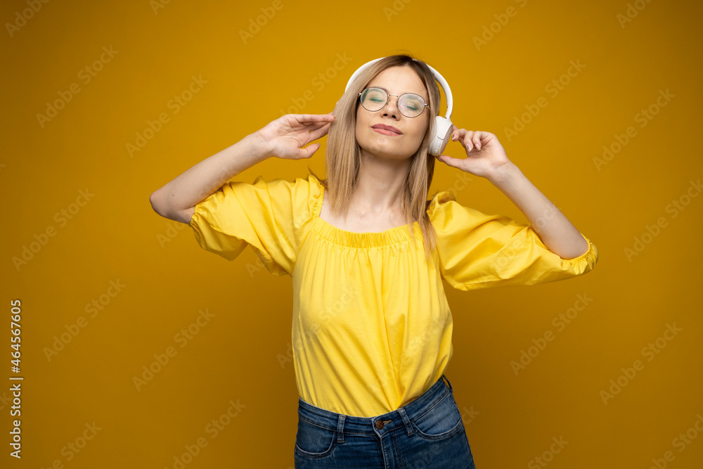 Beautiful attractive young blond woman wearing yellow t-shirt and glasses in white headphones listening music, dancing and laughing on blue background in studio. Relaxing and enjoying. Lifestyle. Beautiful attractive young blond woman wearing yellow t-shirt and glasses in white headphones listening music, dancing and laughing on blue background in studio. Relaxing and enjoying. Lifestyle.