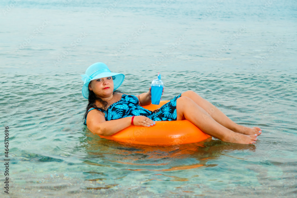 A woman in a straw hat and glasses in the sea in a swimming circle with a cocktail in her hand.