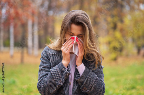 A girl stands on the street in a park in autumn and blows her nose in a handkerchief