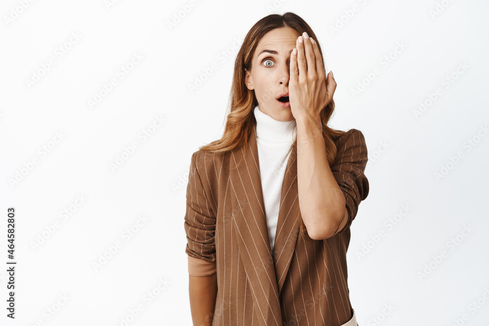Image of corporate woman in suit covers half of face, looking surprised and amazed, saying wow, standing in awe against white background Image of corporate woman in suit covers half of face, looking surprised and amazed, saying wow, standing in awe against white background