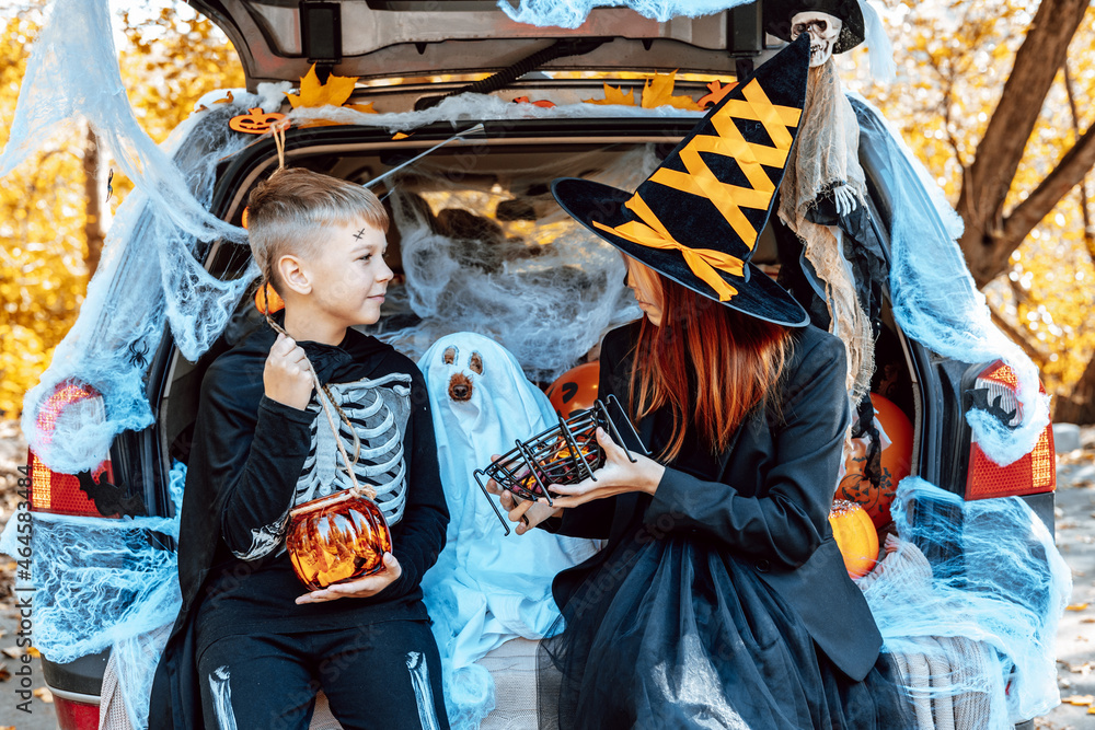 Obraz premium siblings boy in skeleton costume, teenage girl in witch costume and hat and cute poodle dog in ghost costume sits in trunk car decorated for Halloween with web, orange balloons and pumpkins, outdoor