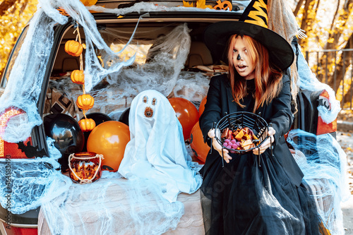 teenage girl in witch costume and hat and cute poodle dog in ghost costume sits in trunk car decorated for Halloween with web, orange balloons and pumpkins, outdoor creative activity concept in autumn