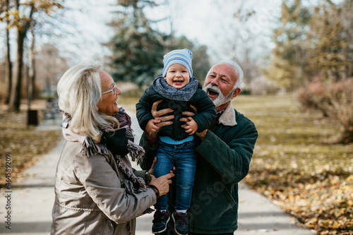 Happy good looking senior couple husband and wife walking and playing with their adorable grandson in public city park