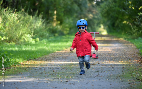 Caucasian child wearing a blue helmet, a red coat and sunglasses, running on a forest path in the fall. A red scooter is in the background and autumn leaves on the ground.
