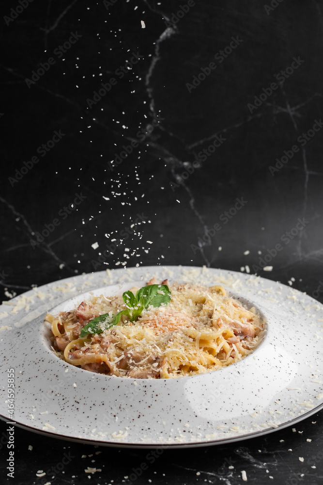 Cheese falling on pasta. Spaghetti with bacon, parmesan, cream sauce and egg yolk on black marble background. Mediterranean gourmet food.