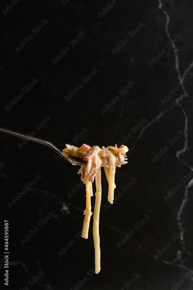 Pasta Carbonara on fork isolated. Spaghetti with bacon, parmesan, cream sauce and egg yolk on black marble background. Mediterranean gourmet food.