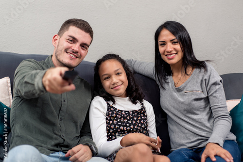 Young interracial family looking at camera from the sofa with the remote control. Concept of family watching TV at home.