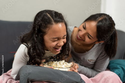 Young latin mother and her daughter laugh on the sofa with popcorn.