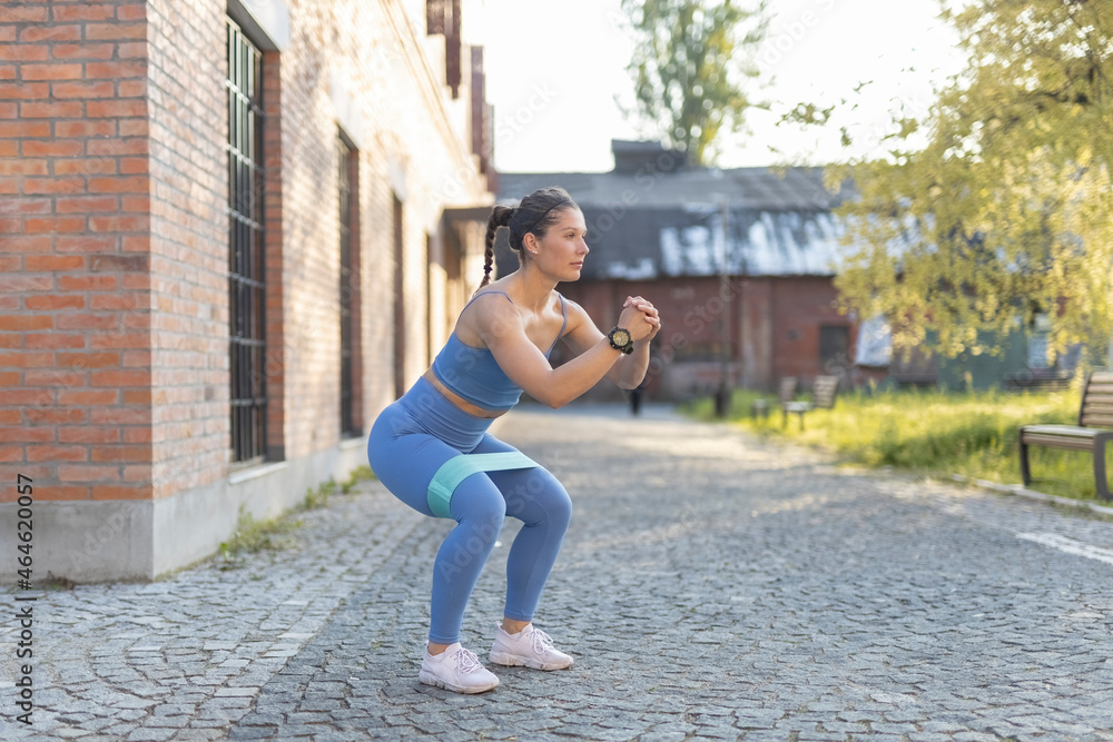 Young fitness female model in blue training sportswear doing leg squat exercises with elastic band in street city valley. Young fitness female model in blue training sportswear doing leg squat exercises with elastic band in street city valley.