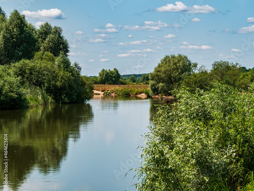 The water surface of the river against the blue sky. Water stream. Lakes and rivers. Coastline. Green leaves of trees. Blue sky. White clouds. Natural landscape. Summer. Reflection in water.
