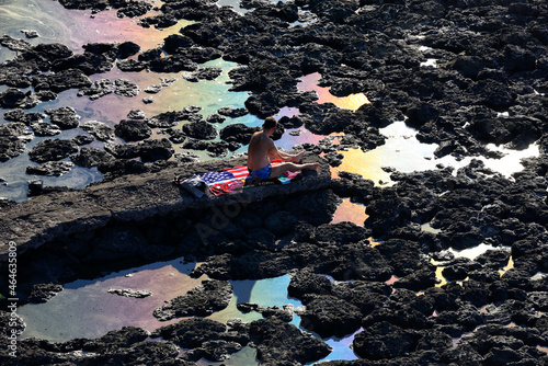 man resting at black volcanic coast formed by lava from Etna volcano, rainbow reflection of crude oil spill on contaminated seawater, Aci Castello, Province of Catania, Sicily, Italy, Europe photo