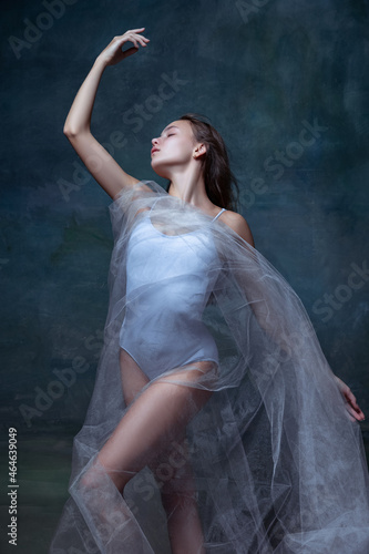 Portrait of young beautiful girl, ballerina posing with white transparent cloth isolated on dark vintage studio background.