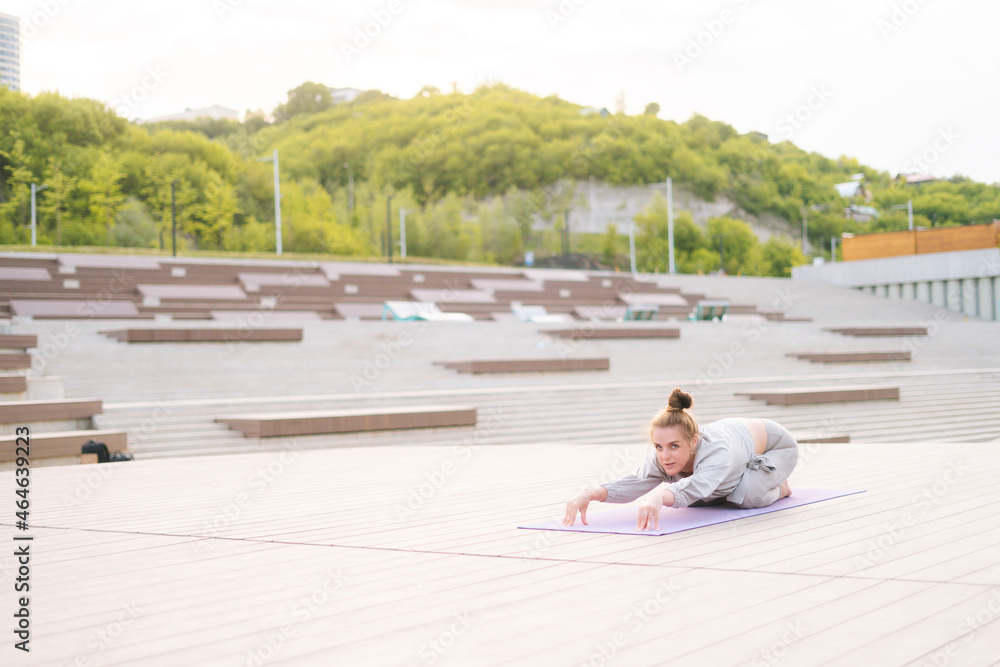Attractive flexible young yogini woman practicing yoga exercisers on fitness mat on summer day outdoors in city park. Calm female with beautiful body in sportswear performing asana pose outside.