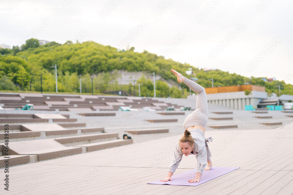 Front view of flexible young yogini woman doing yoga standing on fitness mat on summer day outdoors. Yogini fit sporty female wearing activewear doing stretching exercises at outside.