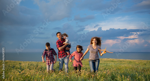 A large family with children launching a toy plane into the sky walking at sunset across a field with a blue sky background with clouds.
