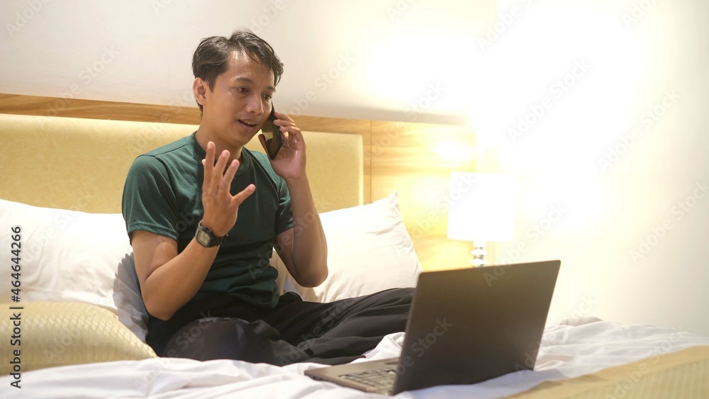 Happy Asian man working with laptop while holding smartphone on bed Happy Asian man working with laptop while holding smartphone on bed