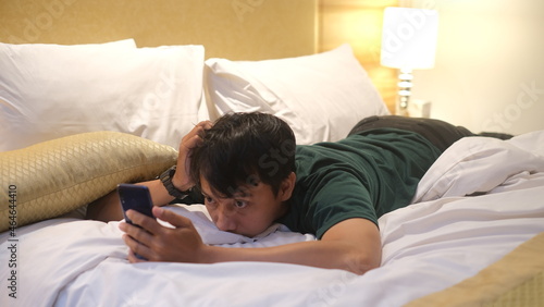 Sleepy Asian man is using smartphone while lying on the bed.