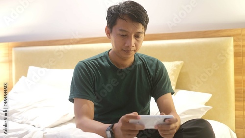 Sleepy Asian man is using smartphone while lying on the bed.