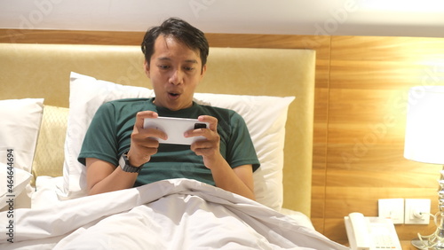 Sleepy Asian man is using smartphone while lying on the bed.